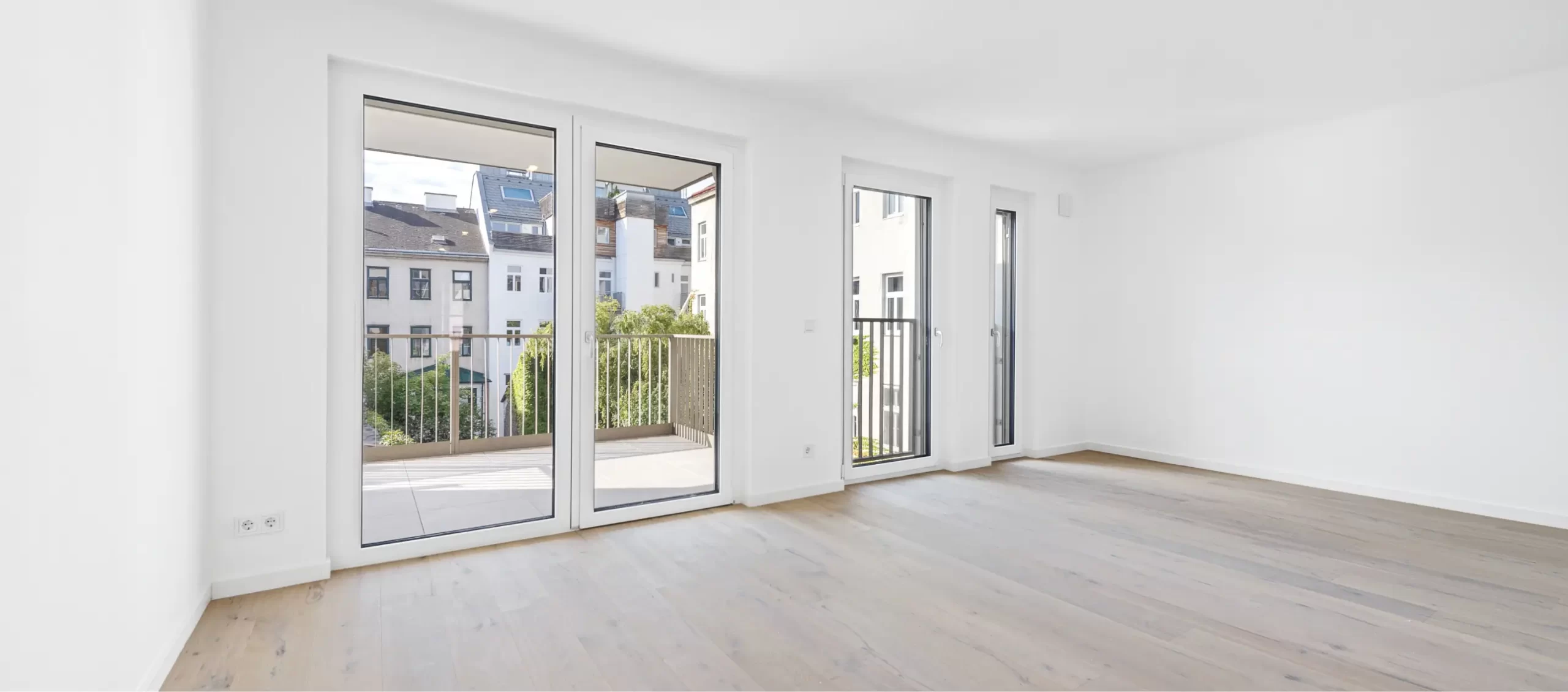 Heller Raum mit Holzboden und großer Balkontür, Blick auf umliegende Gebäude.