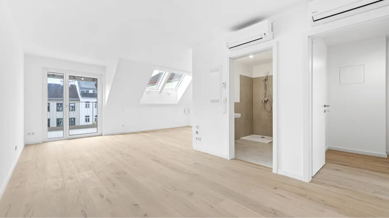 Heller Dachgeschossraum mit Holzboden, Schrägdachfenstern, einer Balkontür und offenem Zugang zu einem Badezimmer.